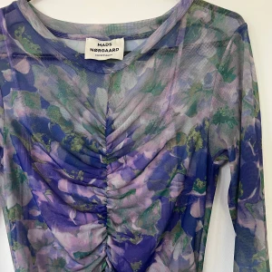 Blommig meshklänning från Mads Nørgaard - Säljer en långärmad, tight meshklänning från Mads Nørgaard med blommigt mönster i lila, grönt och blått. Klänningen har rynkade detaljer framtill och är i genomskinligt material. Perfekt för dig som vill sticka ut med färg och mönster.