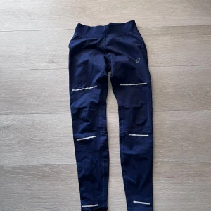 Mörkblå träningstights från Asics - Mörkblå tights från Asics med reflekterande detaljer längs benen. Hög midja och tight passform, perfekta för löpning eller gym. Materialet är stretchigt och andas bra.