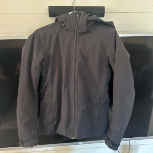 Svart vindjacka från PeakPerformance med huva - Svart vindjacka med huva och dragkedja framtill. Jackan har två fickor med dragkedja och en diskret logotyp på bröstet. Perfekt för blåsiga dagar och enkel att matcha med det mesta.