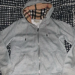 Burberry kofta - Säljer en stilren grå huvtröja från Burberry med dragkedja och klassiskt rutigt mönster i huvan. Tröjan har långa ärmar och en bekväm passform. Perfekt för en avslappnad look. Storlek M passar S