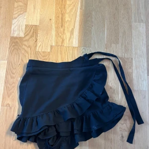Svart omlottkjol med volang - Säljer en svart omlottkjol med volangkant och knytband i midjan. Kjolen har en dragkedja i sidan och är kort i modellen. Perfekt för dig som gillar en feminin och trendig stil. Det är shorts men ser ut som en kjol när man har på sig de