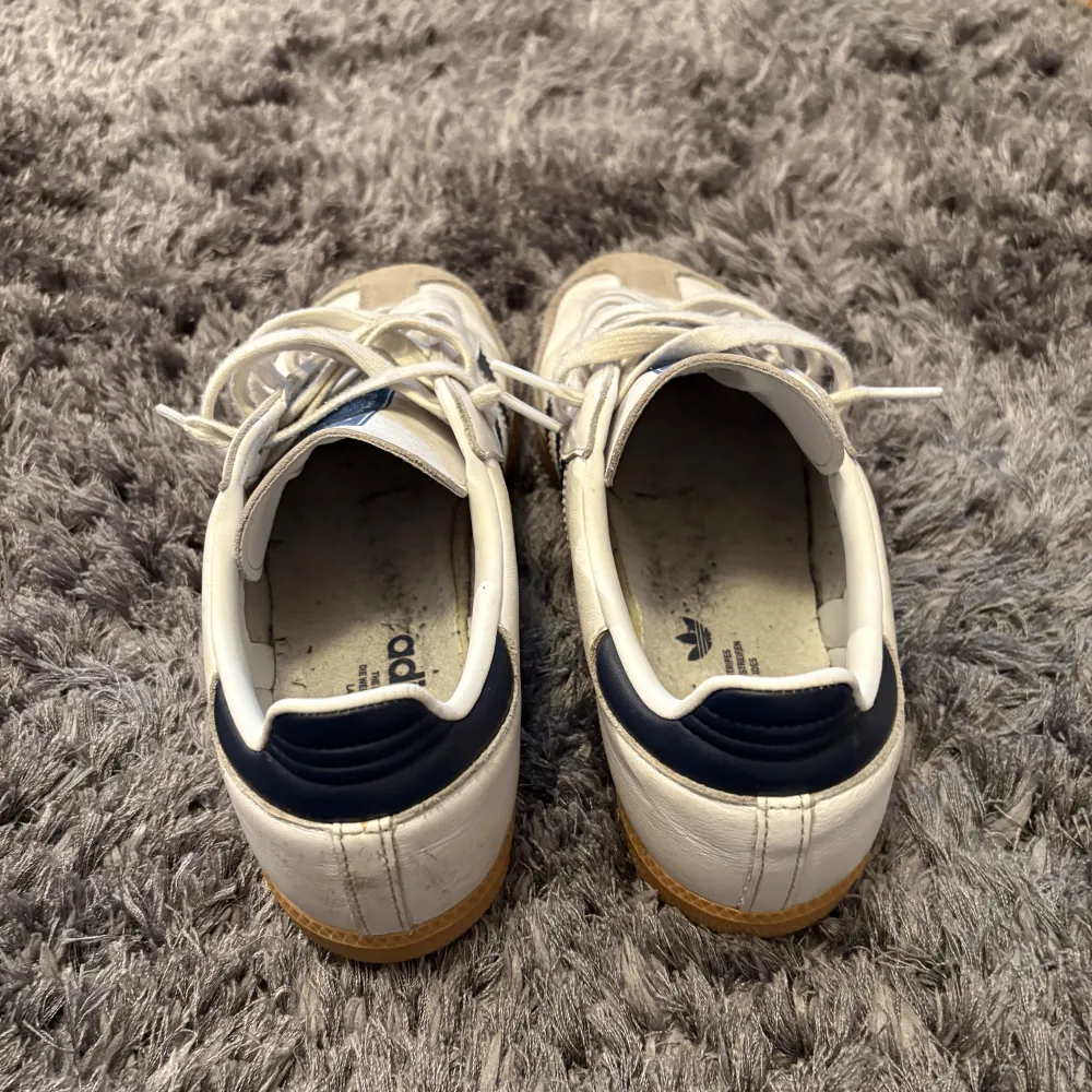 Klassiska Adidas Samba sneakers i vitt med blå ränder och beige mockadetaljer. Skorna har vit ovandel, blå Adidas-logga på plösen och gummisula. De är köpta på Zalando för 1345kr och de är i ett väldigt bra skick👍. Kengät.