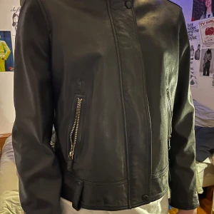 Svart skinnjacka från Massimo Dutti - Säljer en svart skinnjacka från Massimo Dutti i äkta läder. Jackan har lång ärm, ståkrage med tryckknapp och dragkedjor vid fickorna. Snygg och stilren modell som passar till många olika outfits.