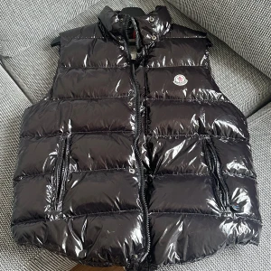 Moncler väst - Exklusiv svart väst med lyxig glans och perfekt passform. Fylld med mjukt material för värme och komfort. Stilsäker look med dragkedjor framtill och på fickor. Perfekt till kalla dagar – både snygg och praktisk.