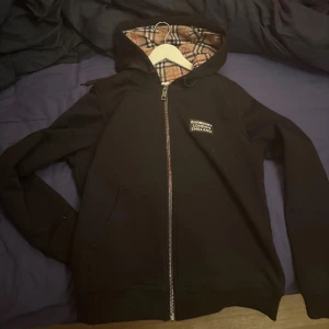 Svart hoodiejacka från Burberry - Svart hoodiejacka från Burberry med dragkedja och klassiskt rutigt foder i huvan. Jackan har fickor framtill och en broderad Burberry-logga på bröstet. Perfekt för dig som gillar stilrena och exklusiva plagg.