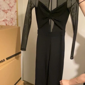 Svart långärmad jumpsuit med meshdetaljer - Säljer en svart jumpsuit med långa meshärmar och mesh upptill. Djup v-ringning med twistad detalj framtill och markerad midja. Byxbenen är raka och långa. Perfekt för en snygg och trendig look.