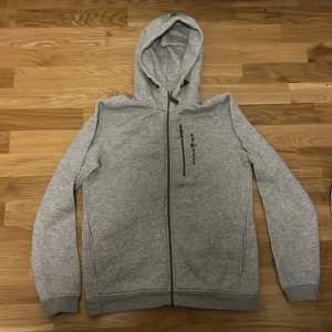 Grå zip hoodie från Sail Racing - Säljer en ljusgrå hoodie från Sail Racing med dragkedja och huva. Tröjan har två fickor framtill och en diskret logga på bröstet. Perfekt för en avslappnad stil och enkel att matcha med det mesta.