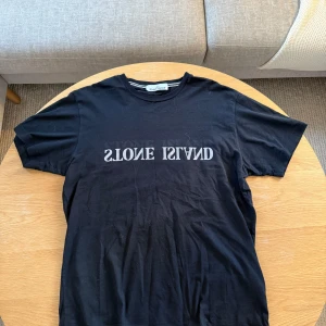 Svart t-shirt från Stone Island - Säljer en svart t-shirt från Stone Island med tryckt logga i vitt på bröstet. Klassisk rund halsringning och korta ärmar. Perfekt för en stilren och avslappnad look.