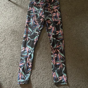 Mönstrade leggings från Stronger - Säljer ett par färgglada leggings från Stronger i storlek S. Byxorna har ett tropiskt mönster i grönt, rosa och blått på svart botten. De är använda fåtal gånger och i bra skick. 