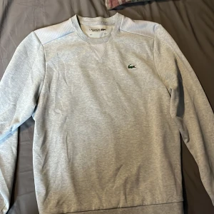 Ljusgrå sweatshirt från Lacoste - Säljer en stilren ljusgrå sweatshirt från Lacoste med klassisk krokodillogga på bröstet. Tröjan har rund halsringning, långa ärmar och ribbade muddar. Perfekt för en avslappnad och sportig look.
