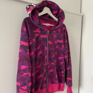Lila och rosa camo zip-hoodie - Säljer en hoodie med dragkedja i lila och rosa camouflage-mönster. Tröjan har huva med tryck, fickor fram och ribbade muddar. Perfekt statement-plagg för streetwear-stilen.
