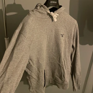 Grå hoodie från GANT med huva - Säljer en grå hoodie från GANT i storlek M. Tröjan har en klassisk huva med snörning, magficka och GANT-logga på bröstet. Mjuk bomullskänsla och långärmad passform, perfekt för chill dagar. Enkel och stilren design som funkar till jeans eller joggers. Hör av er vid funderingar🤗