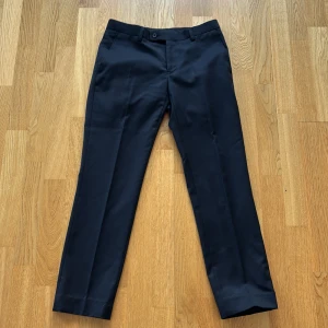 Svarta kostymbyxor Dressmann slim fit - Svarta kostymbyxor från Dressmann i slim fit-modell. Klassisk rak design med bälteshällor, knapp och dragkedja fram samt två bakfickor med knapp. Materialet är mjukt och har en lätt glansig finish som ger en clean look. Perfekt för dig som gillar stilrena och enkla byxor.