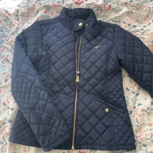 Mörkblå quiltad jacka från Hampton Republic  - Quiltad mörkblå jacka från Hampton Republic med guldfärgade detaljer och dragkedja framtill. Jackan har ståkrage, två fickor med knapp och förstärkta armbågar. Perfekt för nu när vädret börjar bli lite kyligt! 