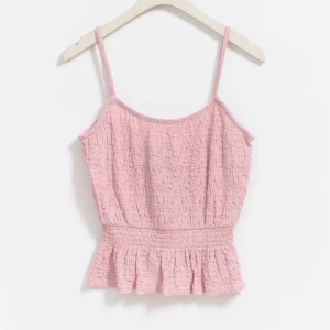 Ljusrosa topp med volang - Supersött ljusrosa linne med smala axelband och volangkant nedtill. Toppen har en smockad midja som ger en snygg form och är tillverkad i ett krinklat bomullstyg. Perfekt för varma dagar och ger en chill vibe till din outfit.