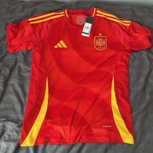 Spaniens herrlandslag fotbollströja Adidas M - Säljer en officiell Spanien fotbollströja från Adidas i rött med gula detaljer och emblem på bröstet. Tröjan har korta ärmar, rund hals och är tillverkad i lätt, ventilerande polyester med Aeroready-teknologi. Snygga gula ränder på axlarna och en gul blomma i nacken. Tröjan passar M och L