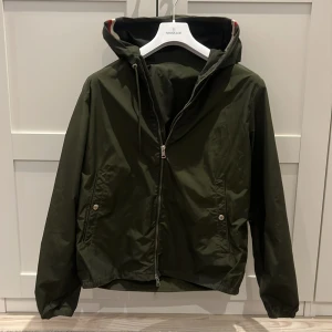 Grön vindjacka från Moncler - Snygg mörkgrön vindjacka från Moncler med huva och coola röd, vit och blå detaljer på kanten. Jackan har dragkedja framtill, två fickor med knappar och Moncler-logga på ärmen. Perfekt för dig som gillar stilren design och vill ha något som sticker ut.