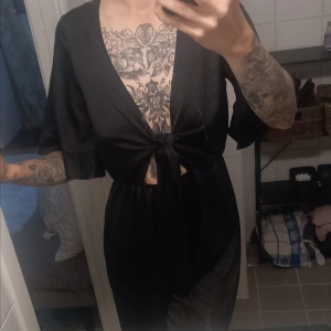 Svart jumpsuit  med knyt - Säljer en svart jumpsuit med djup v-ringning och knyt framtill. har korta, vida ärmar och är tillverkad i ett mjukt, glansigt material som ger en lyxig känsla. Perfekt för dig som gillar en stilren och edgy look.