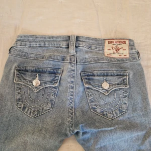 True Religion ljusblå bootcut jeans - Säljer ett par ljusblå jeans från True Religion med klassiska broderade fickor och silverfärgade knappar bak. Modellen har bootcut-ben och låg midja, tillverkade i mjukt jeanstyg. Nya kostar 1300 kr
