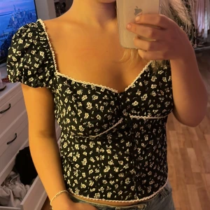 Svart blommig blus med puffärm - Supersöt svart blus med vita små blommor och puffiga korta ärmar. V-ringad med spetskant och knappar framtill. Sitter snyggt med markerad midja och har en romantisk vintage-vibe. Perfekt till jeans eller kjol för en cool look.