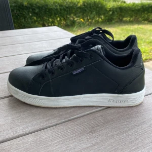 Svarta sneakers från Kappa, strl 38 skor till  - Säljer ett par stilrena svarta sneakers från Kappa i storlek 38. Skorna har en klassisk låg profil med rund tå, snörning och vit platt sula. Ovandelen är i syntetmaterial med diskret Kappa-logga på sidan och bak. Perfekta för dig som gillar enkel och clean streetstil. Nyskick. De är nog mer som storlek 37 men mått kan fås av sulan. 