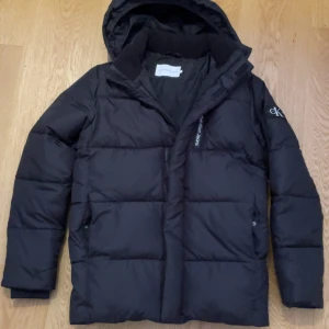 Svart pufferjacka från Calvin Klein  - Svart pufferjacka från Calvin Klein Jeans med huva och diskret logga på ärmen. Jackan har dragkedja framtill och två sidofickor. Den är quiltad och har en clean, modern look som passar perfekt när det är kallt ute.