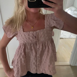 Ljusrosa blus med knappar - Supersöt ljusrosa blus med korta ärmar och fyrkantig ringning. Blusen har en luftig, volangig passform och är gjord i ett strukturerat tyg med knappar framtill. Perfekt för dig som gillar romantiska och chill vibes.