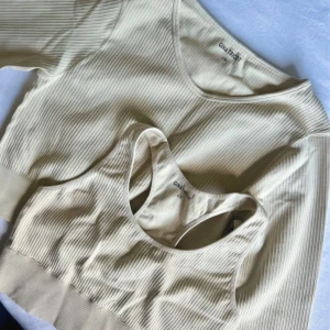 Beige ribbad topp och sport-bh Gina Tricot - Säljer ett set från Gina Tricot med en croppad topp och matchande sport-bh i beige ribbad bomull. Båda har en snygg, enkel design med breda band och rundad hals. Perfekt för chill dagar eller träning. Snyggt att bära som set eller var för sig.