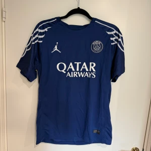 PSG Jordan fotbollströja S - Blå PSG fotbollströja från Jordan med vit logga och tryck. Kortärmad modell i lätt Dri-Fit material som andas. Snygga vita detaljer på axlarna och Paris Saint-Germain emblem på bröstet. Perfekt för dig som vill ha en stilren och sportig look.