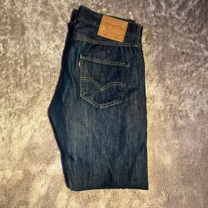 Levi's 501 mörkblå jeans straight fit - Säljer ett par klassiska Levi's 501 jeans i mörkblå tvätt med raka ben och fem fickor. Jeansen har den ikoniska läderpatchen bak i midjan och kontrastsömmar. Perfekt för dig som gillar tidlös stil och vill ha ett par jeans som funkar till allt.