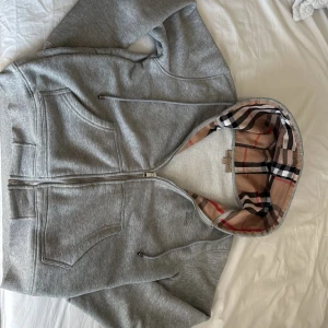 Burberry hoodie🤩 - Säljer en grå hoodie från Burberry med dragkedja och klassisk rutmönstrad insida på huvan. Tröjan har långa ärmar, två fickor fram och snygga Burberry-detaljer på dragkedjan och snörningen. Perfekt för dig som vill ha en stilren och lyxig look. (Har haft den i kanske 2 månader och har blivit för liten på mig är 188 och den sitter lite för kort på mig men annars inget problem. Skriv om ni vill ha andra bilder 😁 💬 )