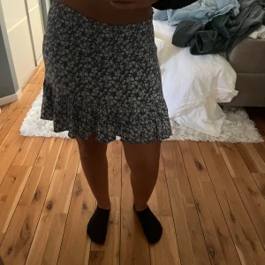 Blommig volangkjol i mörkblått - Supersöt kort kjol med blommigt mönster i vitt på mörkblå botten. Kjolen har volangkant nedtill som ger en extra feminin touch. Perfekt för dig som gillar en avslappnad men ändå trendig stil.