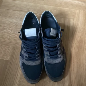 Philippe Model blå-grå sneakers - Snygga sneakers från Philippe Model i en mix av mörkblått och grått med vita detaljer på hälen. Skorna har rund tå, snörning och platt sula. Materialet är en kombination av mocka och syntet, vilket ger en cool och sportig vibe. Perfekta för dig som gillar stilrena och trendiga sneakers. Har något orange färg lite på det vita på baksidan av skon. Perfekt vår/höst sko.