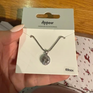 Silverfärgat halsband med glittrig rund berlock - Snyggt halsband från Appear med en rund berlock i silverfärg, dekorerad med gnistrande stenar som ger en glittrig effekt. Kedjan är tunn och elegant, perfekt för att lyfta din outfit med lite extra bling. Passar dig som gillar stilrena och trendiga accessoarer.