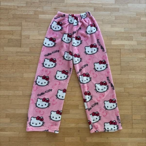 Rosa Hello Kitty fleecebyxor - Supermysiga rosa fleecebyxor med Hello Kitty-tryck över hela byxan. De har elastisk midja och är löst sittande med raka ben. Perfekta för chill hemma eller sleepover med kompisar. Mönstret är gulligt med vita Hello Kitty-ansikten och röda rosetter. Ganska korta i benen så man får ha dem rätt så lågmidjat. Märke är okänt eftersom det inte var jag som köpte dem.
