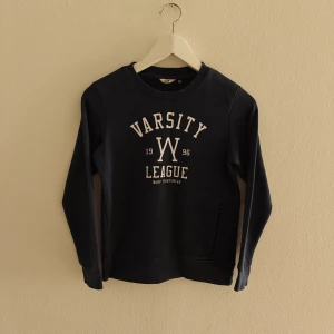 Warp tröja - Mörkblå sweatshirt från Warp med trycket 'Varsity League 1996' i vitt på bröstet. Tröjan har rund hals, lång ärm och en praktisk ficka med dragkedja på sidan. Perfekt för dig som gillar sportig och avslappnad stil.