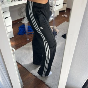 Svarta Adidas med dragkedja - Äkta Adidas med klassiska vita ränder längs sidorna och Adidas-logga på låret. Byxorna har elastisk midja, raka ben och dragkedja vid benslut. Perfekta för chill eller sport, tillverkade i mjuk polyester som andas.