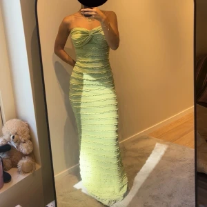 Ljusgrön lång axelbandslös klänning - Säljer en ljusgrön lång klänning med axelbandslös design och snygg twistad detalj vid bysten. Klänningen har ett ribbat, vågigt mönster längs hela plagget och sitter tight mot kroppen för en smickrande siluett. Perfekt för dig som vill sticka ut med en fräsch färg och cool struktur. Xs men passar S
