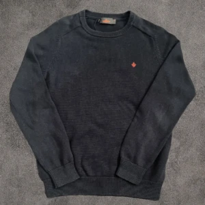 Mörkblå stickad tröja från Morris - Snygg mörkblå stickad tröja från Morris med rund hals och diskret röd broderad logga på bröstet. Tröjan har ribbade muddar vid ärmslut och nederkant, vilket ger en schysst passform. Perfekt för dig som gillar klassisk stil med en modern touch.