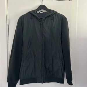 Cardigan från Massimo Dutti - Grön cardigan från Massimo Dutti med huva och dragkedja framtill. Jackan har släta ärmar i stickad material och kroppen i syntet, samt två sidofickor. Perfekt för dig som gillar stilren och enkel design.