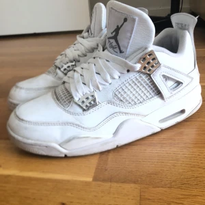 Nike Air Jordan 4 Pure Money sneakers - Nike Air Jordan 4 Pure Money sneakers i helvitt med silverdetaljer. Klassisk high-top siluett med snörning och meshpaneler på sidorna. Skorna har en platt sula och är tillverkade i skinn med coola metalliska accenter. Perfekt för dig som vill ha en clean och stilren look.