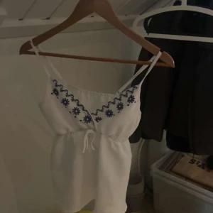 Vit linnetopp med blå broderi - Supersöt vit linnetopp med smala axelband och blå broderade blommor längs v-ringningen. Toppen har knyt i midjan för extra snygg passform och är perfekt för varma dagar. Materialet känns lätt och luftigt, och broderidetaljerna ger en boho-vibe.