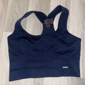 Mörkblå linnetopp från Aim'n - Mörkblå sporttopp Aim'n med breda axelband och racerback-design. Stretchigt material i polyamid och elastan som sitter skönt och ger bra stöd. Perfekt för gymmet eller andra aktiva dagar. Diskret metallmärke framtill för extra stilpoäng. Använd enast 1 gång då den är för liten för mig.