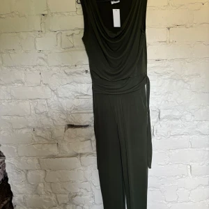 Mörkgrön one peice med drapering - Snygg mörkgrön one peice  med ärmlös design och draperad halsringning. Dressen  har ett knytband i midjan som ger en cool siluett och är tillverkad i ett mjukt, stretchigt material. Perfekt för dig som gillar stilrena och moderna plagg.