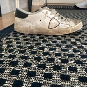 Philippe model skor - Snygga vita sneakers från Philippe model med grå detaljer och svart häl. Skon har snörning, rund tå och platt sula. Materialet är skinn och syntet, vilket ger en klassisk och clean vibe. Perfekt för dig som gillar sportig stil med fransk touch. Har ett ganska stort hål under sulan men de kan man nog tejpa. Det är ett par skit skor har haft dom väldigt länge men vill bara se ifall jag kan få något för dom.