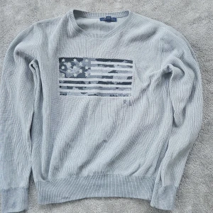 Ljusgrå stickad tröja Polo Ralph Lauren - Snygg ljusgrå stickad tröja från Polo Ralph Lauren med amerikansk flagga i blått och vitt på bröstet. Tröjan har rund hals, ribbade muddar och RL-broderi. Perfekt för dig som gillar klassisk stil med en twist.