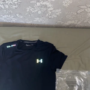 Svart Under Armour t-shirt - Svart Under Armour Threadborne t-shirt med UA-SPEC tryck på axeln och reflekterande logga på bröstet. T-shirten är kortärmad, har en rund hals och är gjord i ett lätt, syntetiskt HeatGear-material som andas. Perfekt för löpning och träning.
