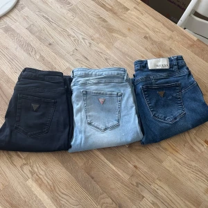 3-pack Guess jeans i olika färger - Säljer tre par jeans från Guess: ett par svarta, ett par ljusblå med slitningar och ett par klassiskt blå. Alla har den ikoniska Guess-loggan på bakfickan och skinny passform. Ljusblåa paret har snygga slitningar framtill för en trendig look. Perfekt för dig som gillar stilrena och moderna jeans.OBS Två paren är lagda på bälteshålare.