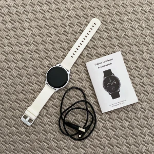 Vit smartwatch från Tobias Jacobsen - Säljer en stilren vit smartwatch från Tobias Jacobsen med rund display och mjukt silikonarmband. Klockan har minimalistisk design och kommer med laddkabel och manual. Perfekt för dig som gillar teknik och vill ha koll på allt direkt på handleden. Obs. Rekommenderar inte om du är nickelallergisk 