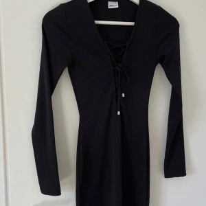 Svart ribbad klänning Gina Tricot - Snygg svart ribbad kortklänning från Gina Tricot i storlek XS. Klänningen har lång ärm, v-ringning med cool snörning framtill och en tight passform som framhäver figuren. Perfekt för dig som gillar stilrena och trendiga plagg.